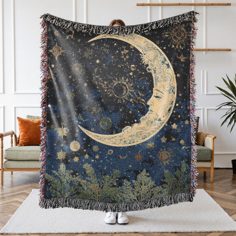 May include: A woven throw blanket featuring a dark blue night sky with a large, golden crescent moon with a human face. The design includes scattered stars, smaller celestial bodies, and green foliage at the bottom edge with fringed borders.