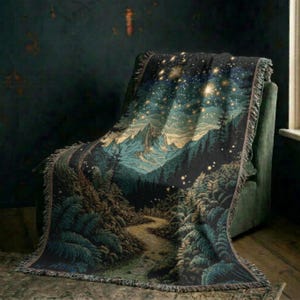 Puede incluir: Una manta tejida que muestra un paisaje de bosque oscuro con un camino sinuoso que conduce hacia montañas nevadas bajo un cielo nocturno estrellado. El textil presenta tonos verde oscuro, azul y dorado con un borde deshilachado.