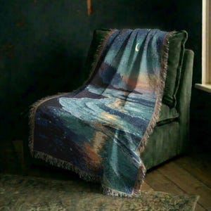 May include: A woven throw blanket featuring a nighttime coastal scene with a crescent moon, a starry sky, and dark blue ocean waves crashing onto a sandy shore. The blanket has a fringed edge and displays a gradient of deep blues, teals, and warm orange.