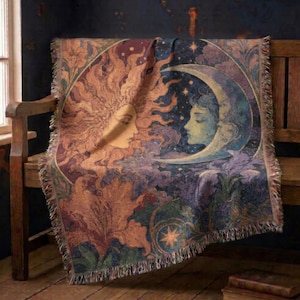 May include: A woven throw blanket featuring a celestial design with a large sun face on the left and a crescent moon face on the right. The tapestry-style fabric displays warm orange and gold tones for the sun and cool blue and purple hues for the moon, surrounded by intricate floral patterns and fringed edges.