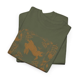 May include: Two olive green t-shirts are stacked. The top shirt features a brown illustration of a rabbit leaping upwards, surrounded by floral and leaf designs. The design is centered on the shirt, creating a nature-inspired theme.