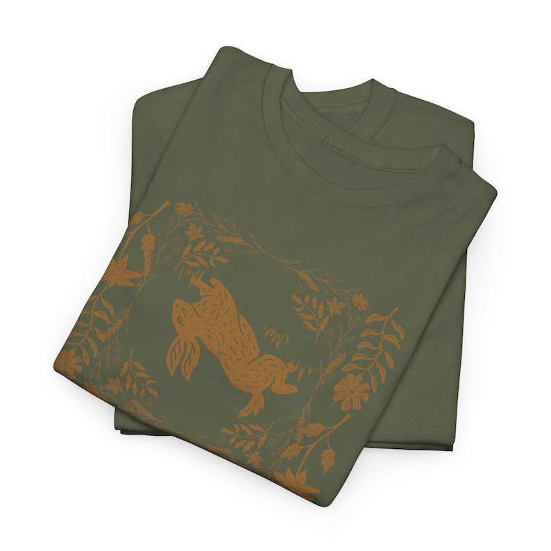 May include: Two olive green t-shirts are stacked. The top shirt features a brown illustration of a rabbit leaping upwards, surrounded by floral and leaf designs. The design is centered on the shirt, creating a nature-inspired theme.