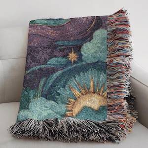 May include: A woven throw blanket featuring a celestial design with a sun, stars, and clouds in shades of teal, purple, and gold. The blanket has a fringed edge with multiple colors. The blanket is folded over a white sofa.
