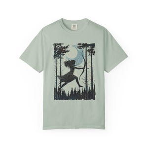 May include: A light green t-shirt featuring a black and blue graphic of a person with a bow and arrow, silhouetted against a forest and moon. The design is centered on the shirt, with a crew neck and short sleeves.