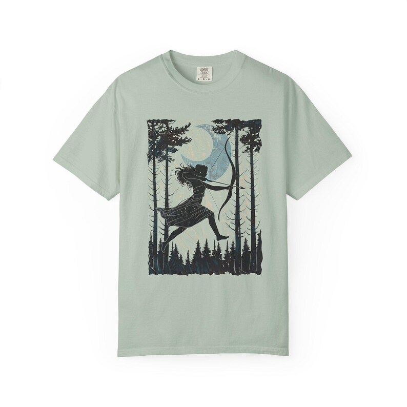 May include: A light green t-shirt featuring a black and blue graphic of a person with a bow and arrow, silhouetted against a forest and moon. The design is centered on the shirt, with a crew neck and short sleeves.