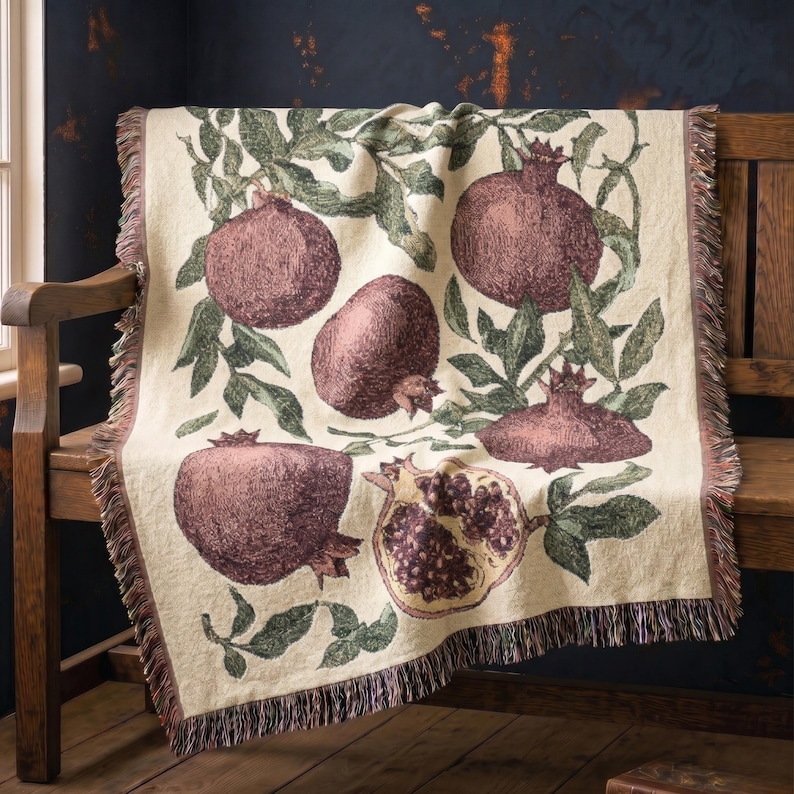 May include: A cream-colored woven throw blanket featuring a repeating botanical pattern of dark red pomegranates and green leaves. The design includes whole pomegranates and one halved fruit showing seeds, finished with a fringed edge on all sides.