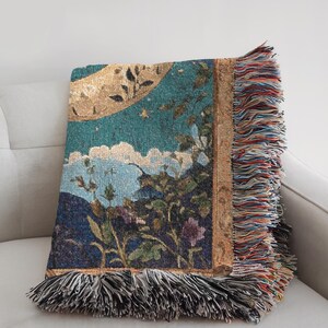 Puede incluir: Una manta tejida que presenta una escena de paisaje con una gran luna dorada, un cielo verde azulado y monta&ntilde;as azul oscuro. El dise&ntilde;o incluye elementos florales y un borde decorativo, acabado con largos flecos multicolores en los bordes.