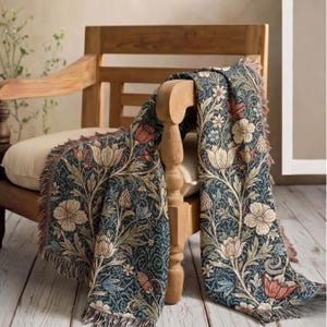 May include: A woven throw blanket featuring a dark blue background with a repeating floral pattern of cream, red, and light blue flowers and leaves. The blanket has a textured appearance with fringed edges draped over a wooden chair.