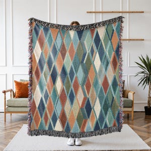 May include: Woven throw blanket with a geometric diamond pattern in shades of blue, orange, green, and cream. The blanket has a fringed edge and is displayed indoors.
