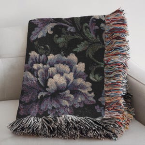 May include: A decorative throw blanket with a floral design. The blanket features a large, detailed flower in shades of purple and gray against a black background. The edges are finished with fringe in multiple colors.