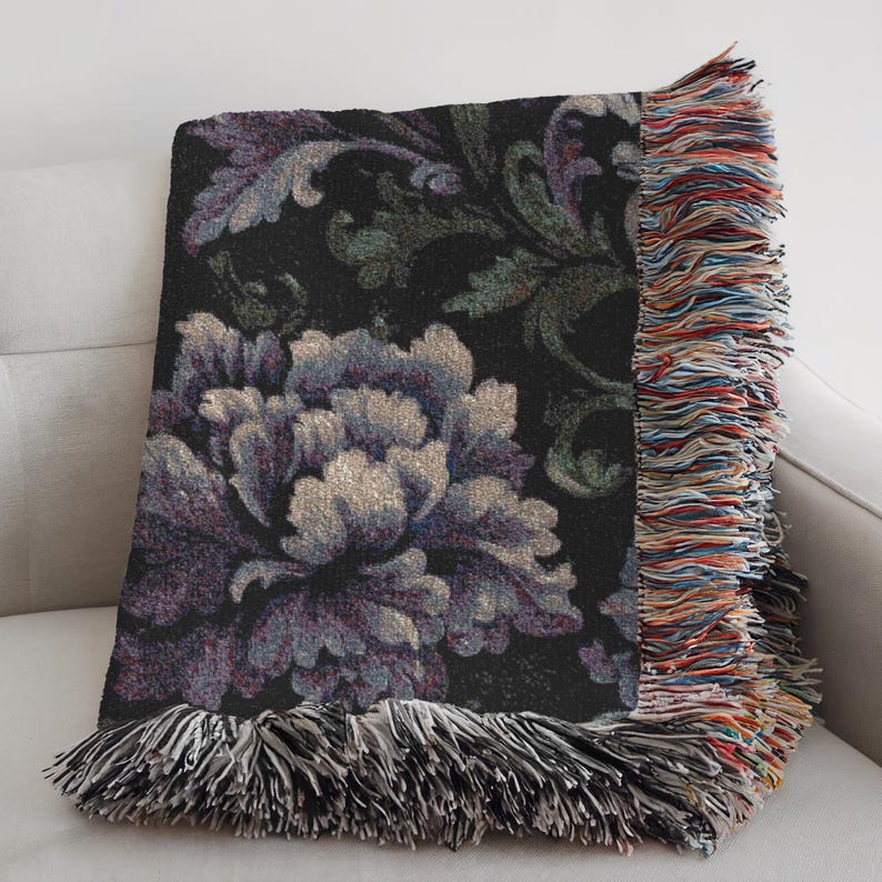 May include: A decorative throw blanket with a floral design. The blanket features a large, detailed flower in shades of purple and gray against a black background. The edges are finished with fringe in multiple colors.