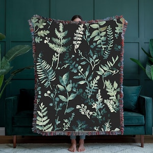 Può includere: Una coperta nera con un motivo botanico di foglie e rami verdi e bianchi. La coperta ha un bordo sfrangiato con fili rossi, blu e verdi. La coperta è esposta davanti a una parete verde scuro e un divano verde.