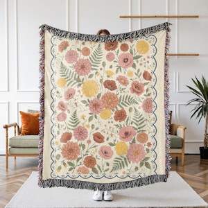 May include: Woven floral throw blanket featuring a cream background with a pattern of pink, yellow, and red flowers and green leaves. The blanket has a scalloped border with a dark blue trim and black fringe.