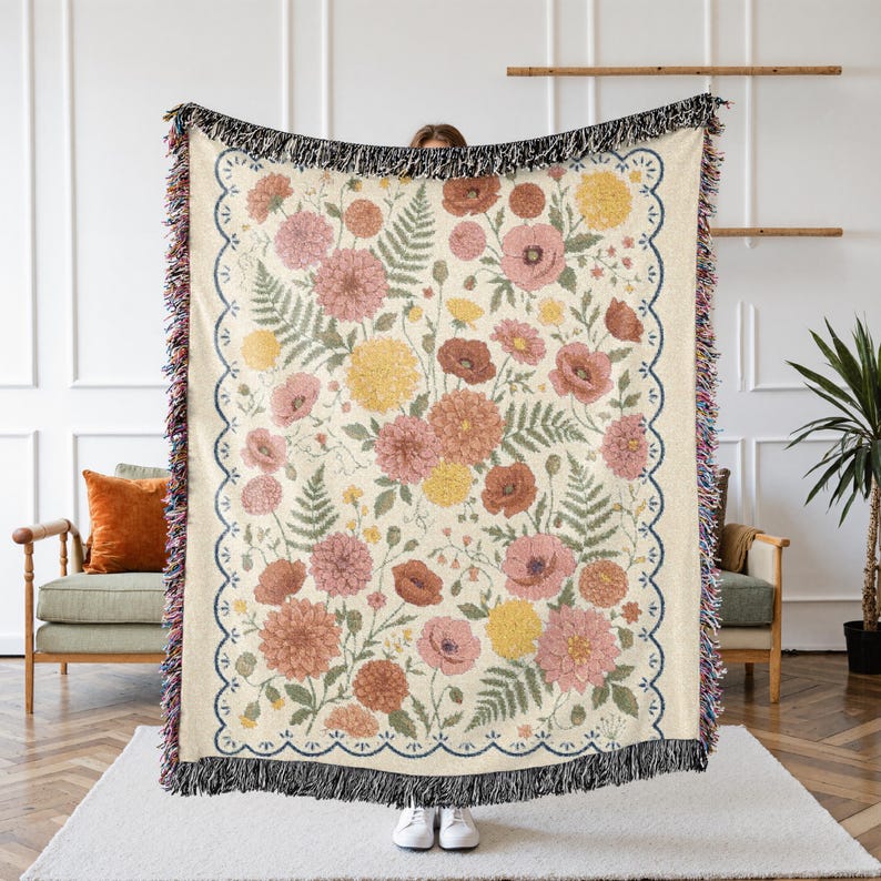 May include: Woven floral throw blanket featuring a cream background with a pattern of pink, yellow, and red flowers and green leaves. The blanket has a scalloped border with a dark blue trim and black fringe.