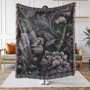 May include: A dark-colored throw blanket with a floral pattern in shades of purple, green, and cream. The blanket has a fringed edge and is held up by a person. The floral design features large, stylized flowers and leaves.