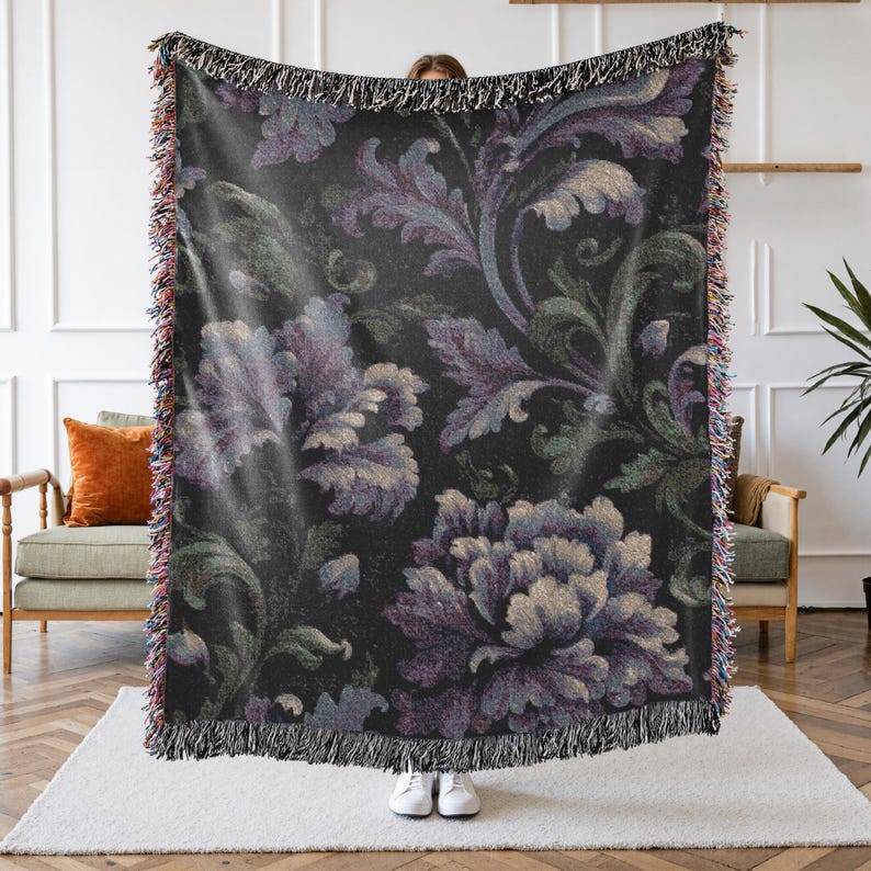 May include: A dark-colored throw blanket with a floral pattern in shades of purple, green, and cream. The blanket has a fringed edge and is held up by a person. The floral design features large, stylized flowers and leaves.