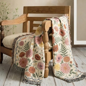 May include: A floral patterned throw blanket draped over a wooden chair. The blanket features a cream background with pink, orange, and yellow flowers, green leaves, and a scalloped edge with fringe.