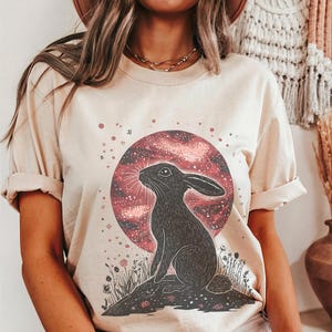 May include: A beige t-shirt with a graphic of a black rabbit silhouetted against a red and pink galaxy-like moon. The moon is surrounded by small white stars.