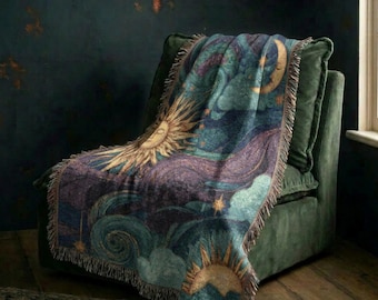 May include: A woven throw blanket featuring a celestial design with a golden sun, a crescent moon, and swirling clouds in shades of deep blue, purple, and teal. The blanket has a fringed edge and is draped over a dark green upholstered armchair.