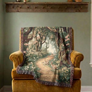 May include: A woven throw blanket featuring a detailed forest scene with a winding dirt path, lush green trees, pink floral accents, and several rabbits. The blanket has a textured fringe border along the edges and is draped over a mustard yellow chair.
