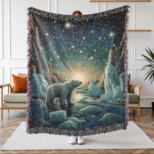 May include: A woven throw blanket featuring a polar bear standing in icy waters beneath a bright sunburst and a starry night sky. The blanket has a dark blue and white color palette with fringed edges on the top and bottom, showcasing a detailed landscape.