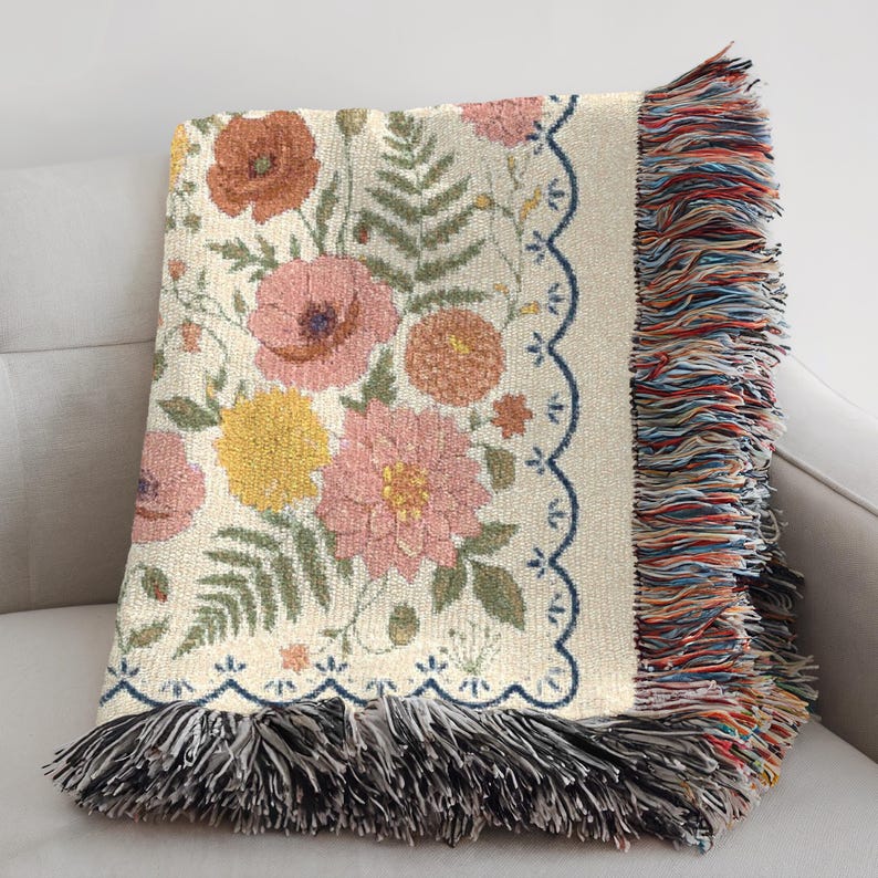 May include: Cream-colored floral throw blanket featuring a repeating pattern of pink, yellow, and orange flowers with green foliage. The blanket has a scalloped edge with a blue border and colorful fringe.