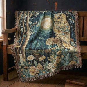 May include: A woven throw blanket featuring a detailed nocturnal scene with a large owl perched on a branch, a full moon, tall trees, and blooming flowers in shades of gold, cream, and deep blue, finished with a decorative fringed edge.