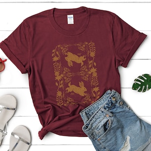 May include: A maroon t-shirt featuring a gold illustration of two rabbits within a floral frame. The shirt is paired with denim shorts and sandals on a white wooden surface. The Gildan tag is visible.