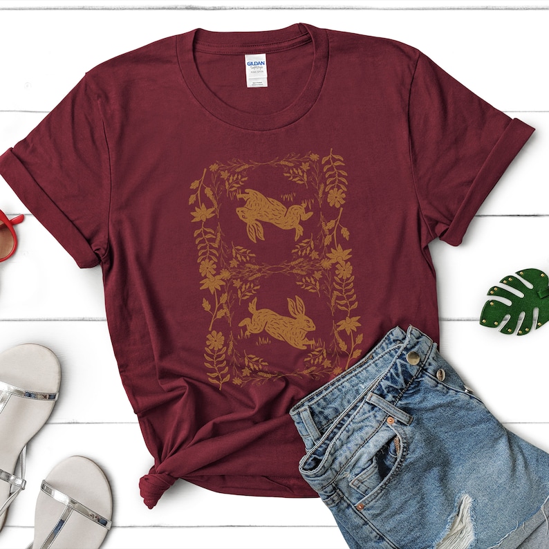 May include: A maroon t-shirt featuring a gold illustration of two rabbits within a floral frame. The shirt is paired with denim shorts and sandals on a white wooden surface. The Gildan tag is visible.