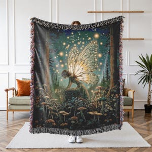 May include: A woven throw blanket featuring a fairy in a forest scene with mushrooms and glowing lights. The fairy has large, detailed wings. The blanket has a black background with a fringed edge.