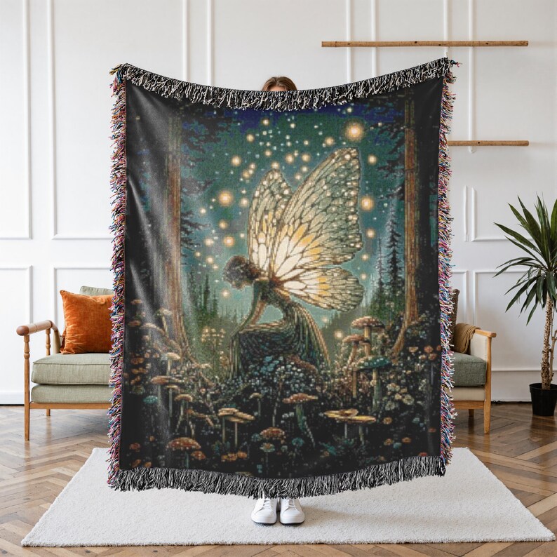May include: A woven throw blanket featuring a fairy in a forest scene with mushrooms and glowing lights. The fairy has large, detailed wings. The blanket has a black background with a fringed edge.