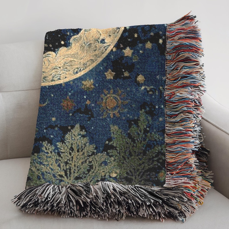 May include: A woven throw blanket featuring a dark blue background with a large golden crescent moon, various stars, and sun motifs. The lower section displays green tree silhouettes. The edges are finished with multi-colored fringe trim.