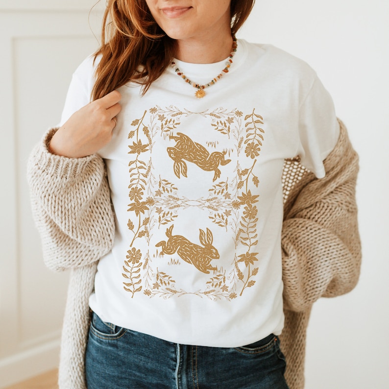 May include: White t-shirt featuring a gold-colored design of two rabbits within a floral frame. The rabbits are depicted in a running pose. The shirt is made of a soft material and is suitable for casual wear.