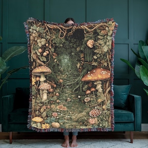 May include: A woven throw blanket featuring a detailed forest scene with various mushrooms, tangled tree roots, and dense foliage in shades of green, brown, and orange. The blanket has a decorative fringed edge on all sides with a multi-coloured border.