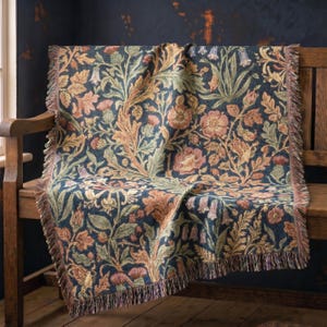 May include: A woven throw blanket featuring a dark navy blue background with a dense botanical pattern of muted orange flowers, green leaves, and thistle-like plants. The rectangular textile has a decorative fringe along the vertical edges.