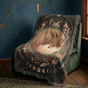 May include: A woven throw blanket featuring a central illustration of a sleeping fox curled in a circle. The design includes surrounding forest elements like mushrooms, ferns, and a lunar moon phase cycle along the bottom edge with fringe trim.