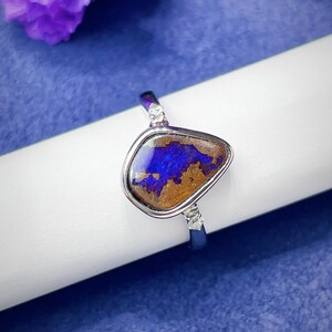 May include: A silver ring featuring a central, irregular-shaped boulder opal with deep blue and brown tones. The stone is set in a smooth metal bezel, flanked by two small, clear, round-cut accent stones on each side of the polished silver band.