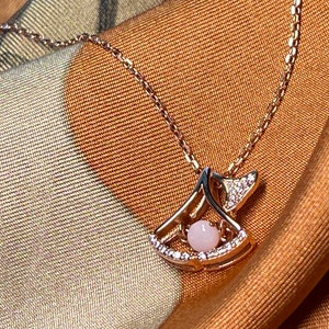 Op de afbeelding: Een delicate gouden ketting met een hanger in de vorm van een gestileerde vleugel met een roze edelsteen in het midden. De hanger is versierd met kleine heldere stenen.