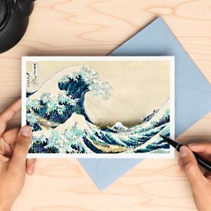 Può includere: Un biglietto di auguri con l'iconica opera d'arte giapponese "La grande onda di Kanagawa" è tenuto sopra una busta blu. Una teiera e una penna nere sono anche sulla superficie di legno. È presente anche un ornamento in ceramica a forma di onda.