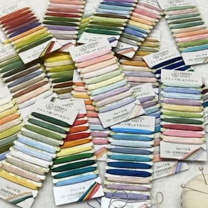 Peut inclure: Plusieurs échevettes de fil à broder de couleurs variées, disposées sur de petites cartes en carton. Les couleurs des fils vont des rouges et oranges vifs aux bleus et verts doux. Accessoires : ciseaux, coussin à épingles et fleurs séchées.