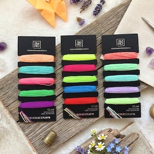 May include: A craft-themed still life with three sets of embroidery floss in vibrant colours. The image also includes gold scissors, a thread spool, needles, and decorative crystals. A paper origami butterfly and lavender sprigs enhance the scene's aesthetic appeal.