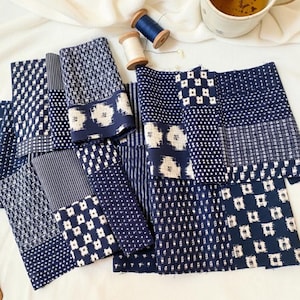 Op de afbeelding: Een assortiment van indigo blauwe en witte stoffen met patronen, met verschillende geometrische en bloemmotieven. De stoffen zijn gerangschikt op een wit oppervlak, met een notitieboek, pen en een kopje thee op de achtergrond.