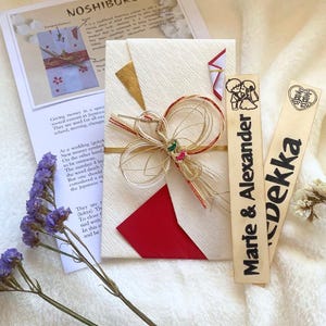 May include: A wedding gift set featuring a white envelope with a red and gold design, a decorative knot, and wooden name tags. The set includes a paper with Japanese text and purple flowers.