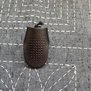 May include: A black leather thimble with a perforated design. The thimble is on a grey fabric background.