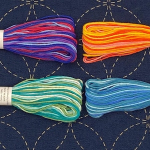 Può includere: Quattro rocchetti di filo da ricamo in diverse combinazioni di colori. I colori sono un mix di rosso, viola, blu, arancione, giallo, verde e verde acqua. I rocchetti sono etichettati con "Cotton 100%" e "Made in Japan".