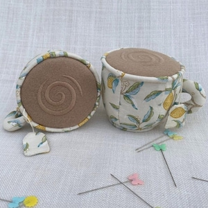 Puede incluir: Una taza de té y un platillo de cerámica blanca con un diseño de impresión de limón. La taza de té está llena de tela marrón y tiene una tapa de tela marrón. El platillo tiene un fondo de tela marrón. Hay varios alfileres de colores esparcidos alrededor de la taza de té y el platillo.