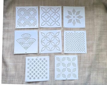 May include: A collection of seven white stencils with various geometric and stylized designs. The stencils feature patterns such as waves, circles, stars, fans, and floral motifs. Each stencil is square and has small holes in the corners.