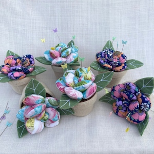 May include: Six fabric flower pincushions in pots. The flowers are made of colourful floral patterned fabric and are arranged in a cluster. The pots are brown and have green leaves.