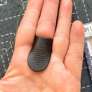 Puede incluir: Un dedal de cuero negro con un patrón de cuadrícula en la parte superior. El dedal se lleva en el dedo índice de una mano. La imagen también muestra un paquete de agujas Sashiko con el texto "Sashiko Needle" y "大 小 各 2 本 針" en japonés.