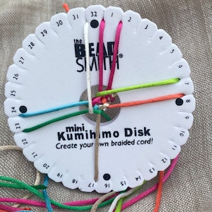May include: A white plastic Kumihimo disk with a black dot at the centre and numbers 1 to 32 around the edge. The disk has the text "the Bead Smith" and "mini. Kumihimo Disk Create your own braided cord!" printed on it. Five strands of colourful cord are threaded through the disk, creating a braided pattern.